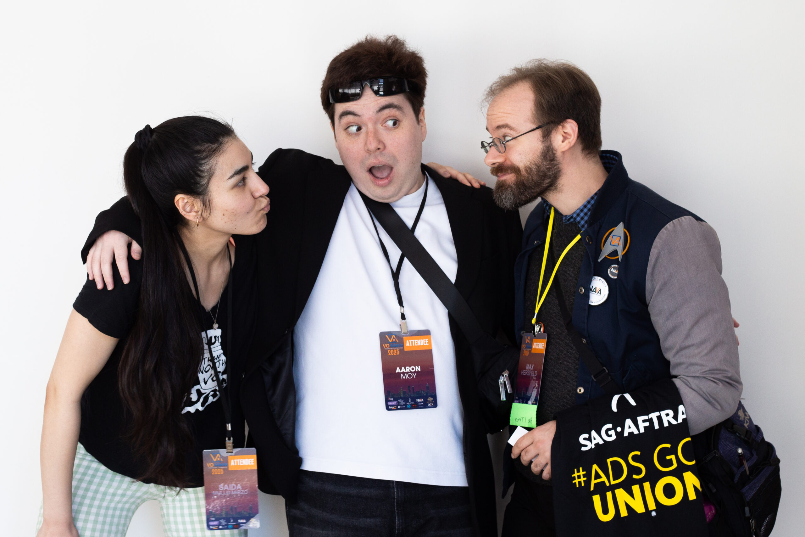 Three attendees at VO Atlanta 2025 stand closely together smiling and making playful faces. The person in the middle has a surprised expression, with one arm around each of the others. All wear conference badges, and the person on the right carries a SAG-AFTRA tote bag with "#AdsGoUnion" written on it.