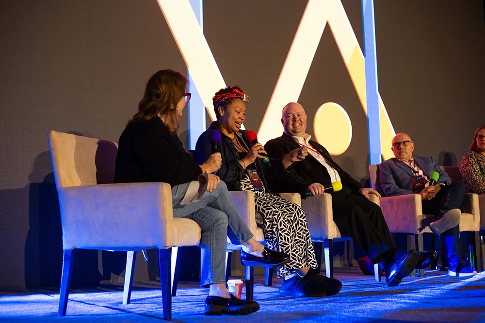 A panel of voice-over professionals speak and laugh during a main stage discussion at VO Atlanta 2025. One woman speaks into a red microphone, surrounded by fellow panelists holding microphones in various colors. The large VO Atlanta logo is visible on the screen behind them.