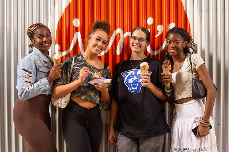 Four Young Women Stand Together In Front Of A Corrugated Metal Wall With An Orange And White Logo. 