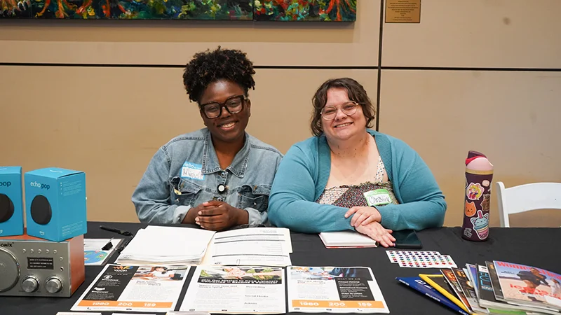 Two Women Sit Behind A Table At An Event, Smiling. The Table Is Covered With Informational Materials, A Radio, And Two Blue Devices.