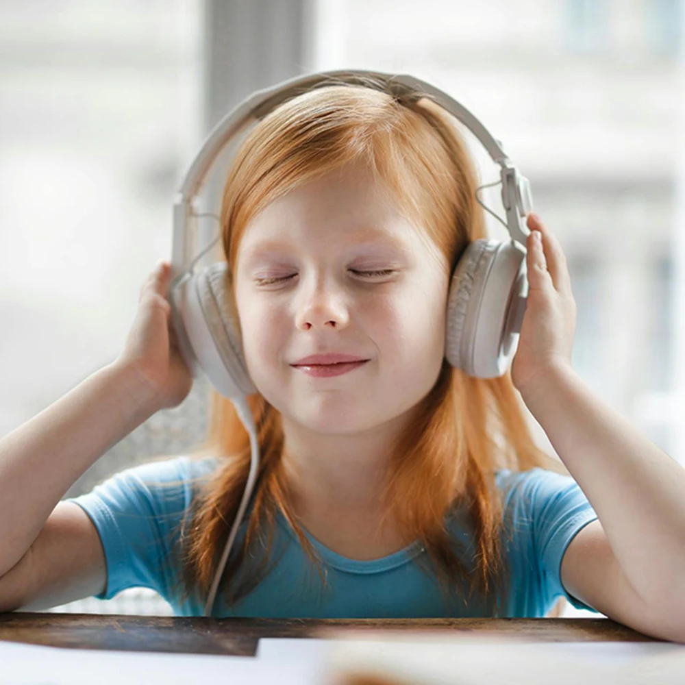 A young girl with red hair in braids sits at a wooden table, eyes closed and smiling gently, holding large white headphones to her ears.