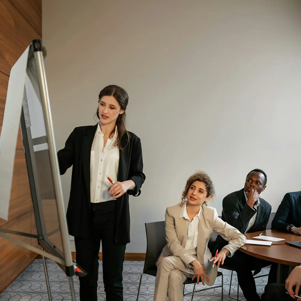 A woman in a black blazer stands presenting at a flip chart, gesturing as she speaks, while colleagues seated around a round conference table listen attentively in a modern meeting room.