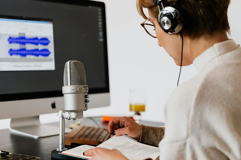 Person wearing headphones records audio at a desk with a microphone, computer displaying audio waveforms, and an open book.